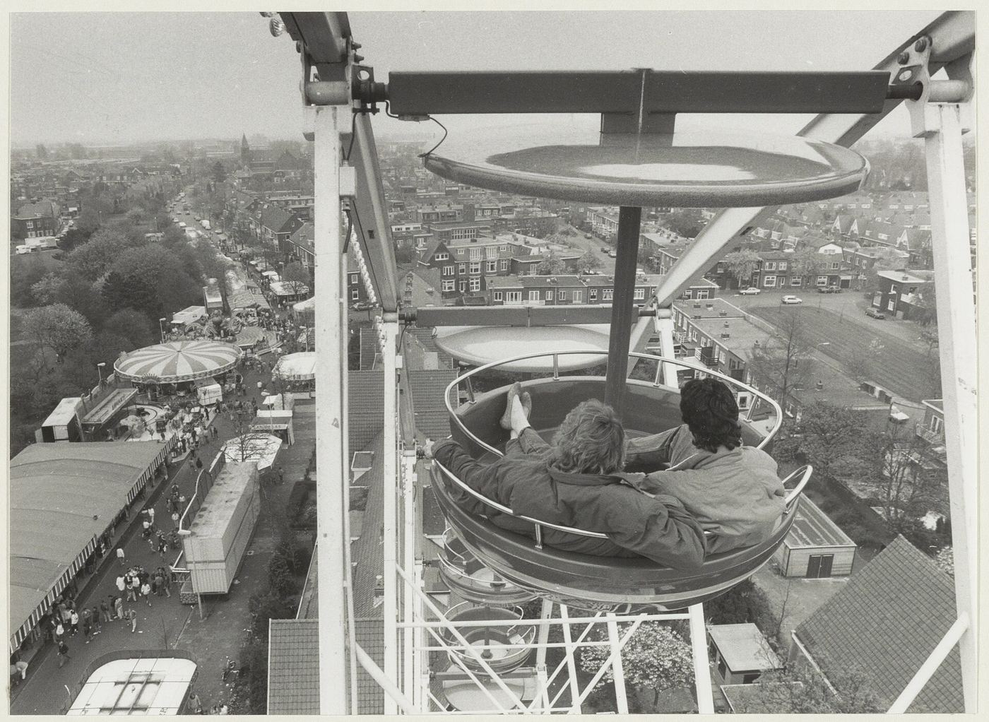 Zwart-witfoto van twee kermisbezoekers in een reuzenrad, uitkijkend over de kermis en de stad